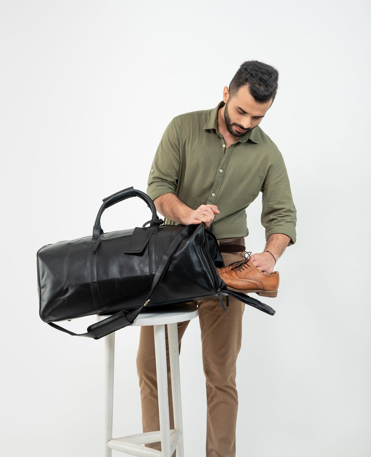 May include: A black leather duffel bag with a brown leather handle and a zipper closure. The bag is being packed with a brown leather shoe.
