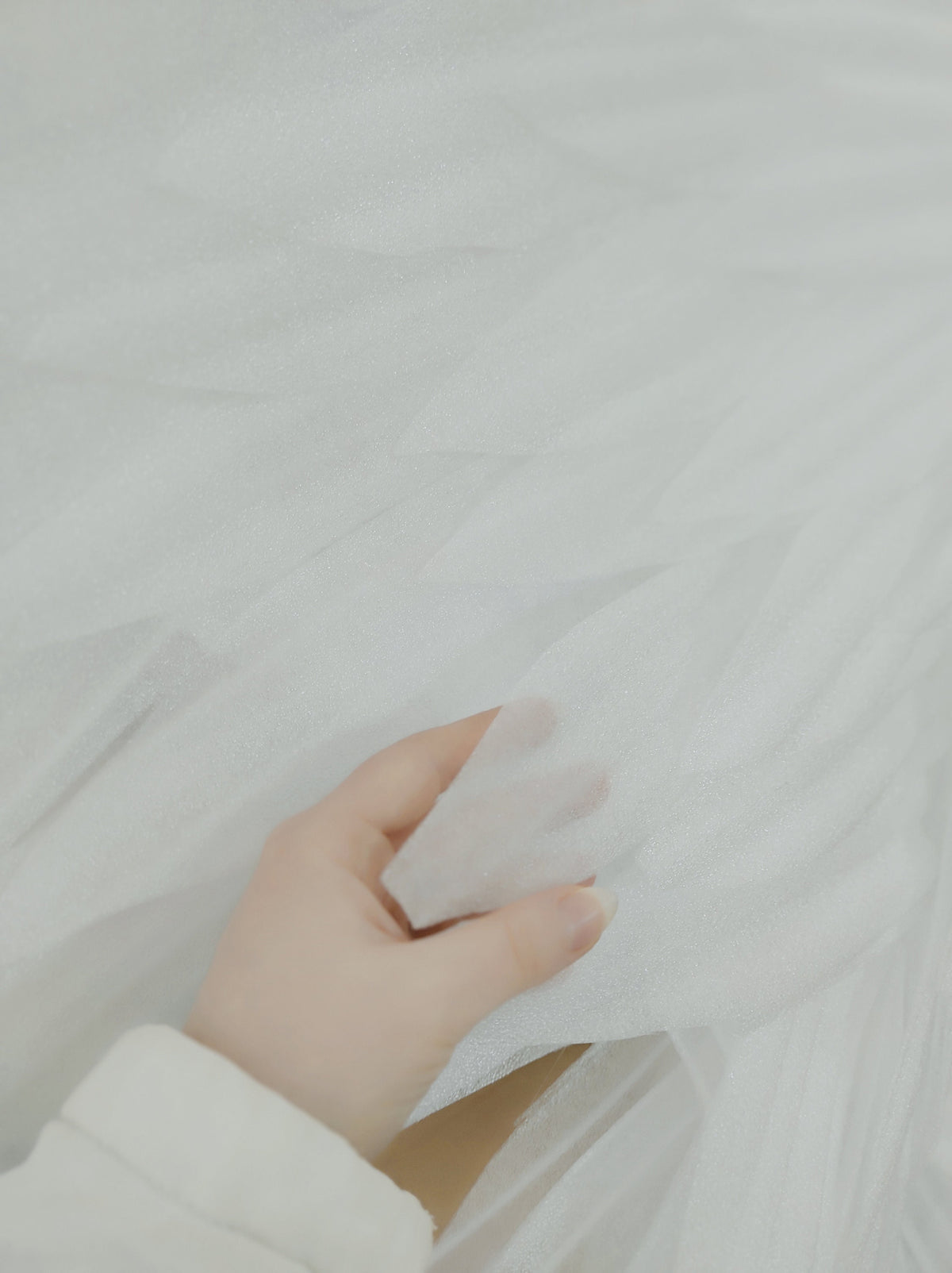 May include: A hand holding a piece of white tulle fabric. The fabric is soft and delicate, with a subtle shimmer.