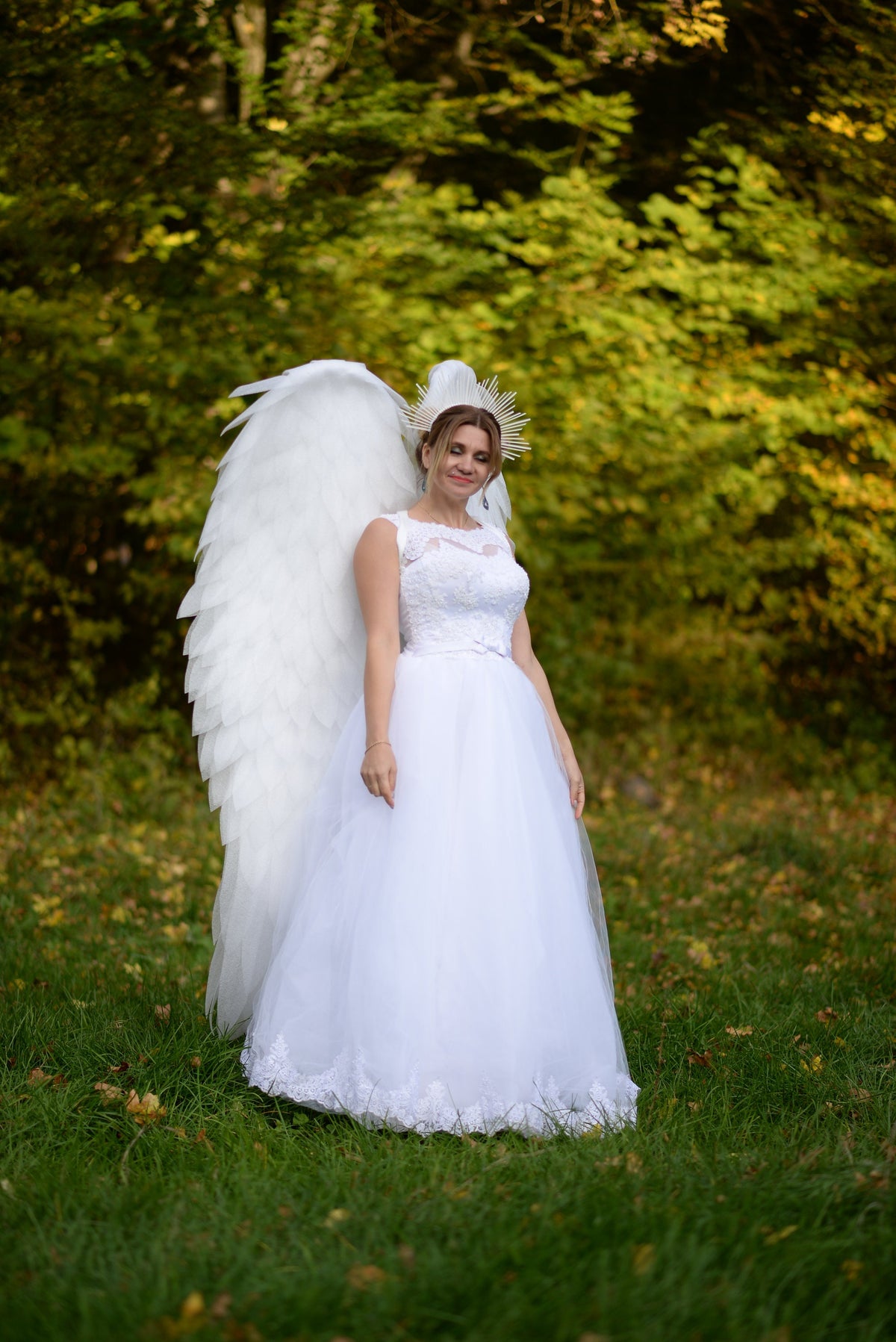 May include: A woman wearing a white wedding dress with a white feathered angel wing and a gold crown stands in a grassy field.