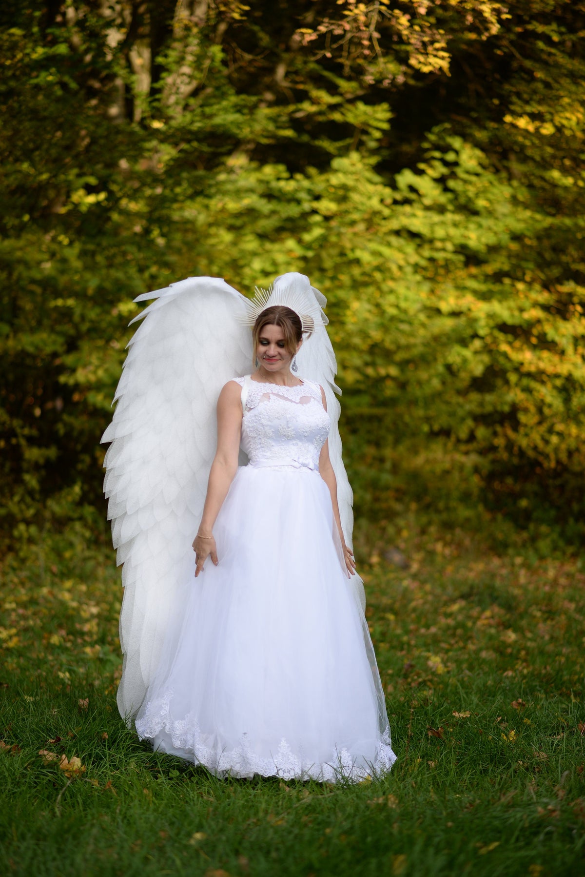 May include: A woman wearing a white wedding dress with a lace bodice and a white feathered angel wing costume. She is standing in a grassy field with fall foliage in the background.