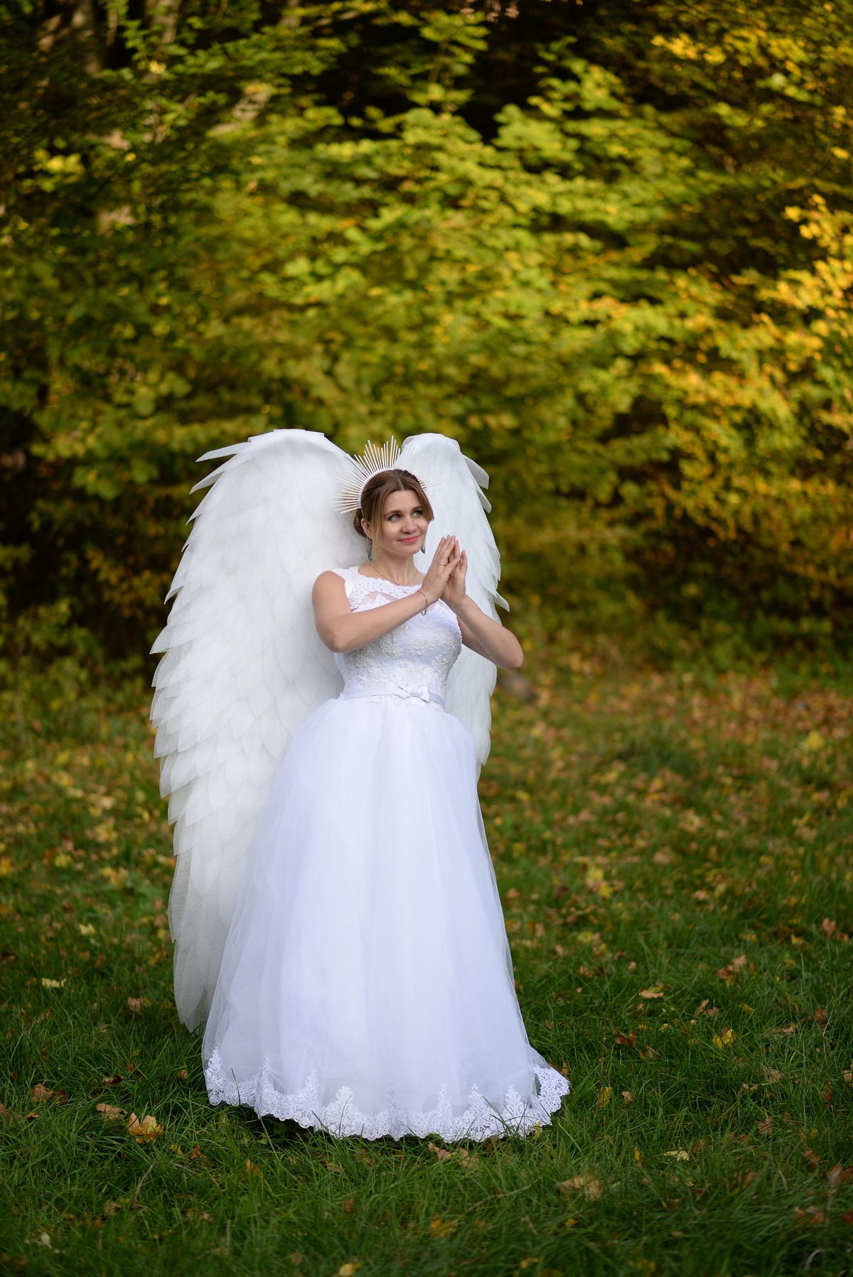 May include: A woman wearing a white wedding dress and a gold crown stands in a grassy field with large white angel wings. She is holding her hands together in front of her chest.