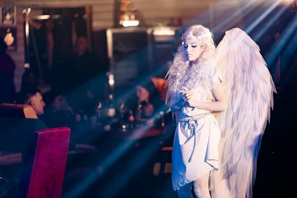 May include: A woman wearing a white dress and angel wings stands in a dimly lit room with a spotlight shining on her. She has long white hair and is holding her hands together in front of her.