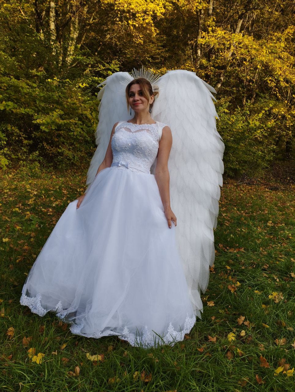 May include: A woman wearing a white wedding dress with a lace trim and a white feathered angel wing costume. She is standing in a grassy area with fallen leaves.