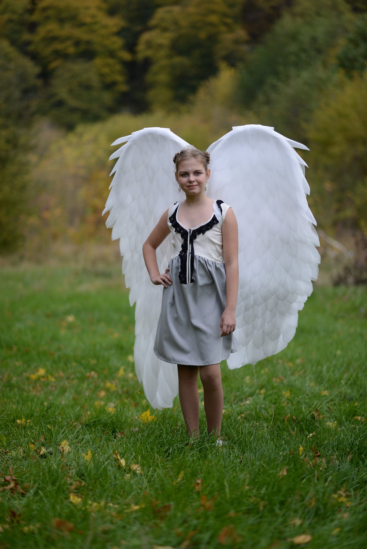 May include: A young person wearing a gray dress with black lace trim and white angel wings. The person is standing in a grassy field.