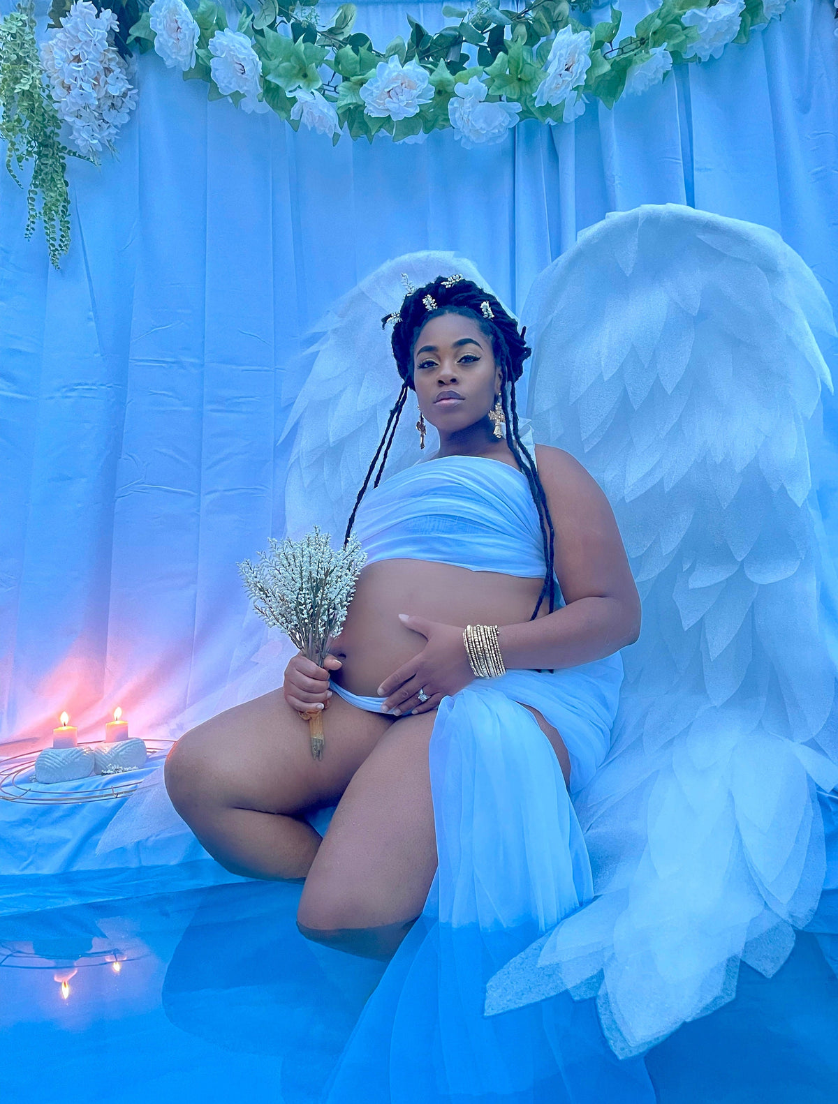 May include: A pregnant woman wearing a white dress and angel wings sits in a pool of water. She is holding a bouquet of white flowers and has her hand on her stomach.