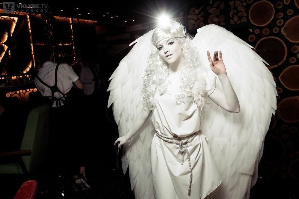 May include: A woman dressed as an angel with white wings and a white dress. She is standing in a dimly lit room with a wooden wall behind her.