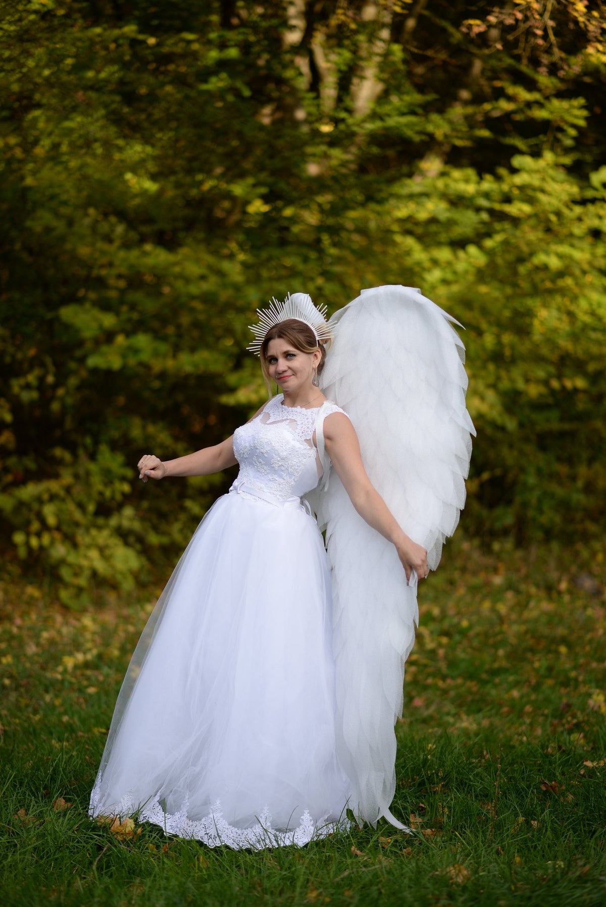 May include: A woman in a white wedding dress with a lace bodice and a full skirt, adorned with large white feathered angel wings. She wears a halo-like headpiece, posing in a natural outdoor setting with green foliage.