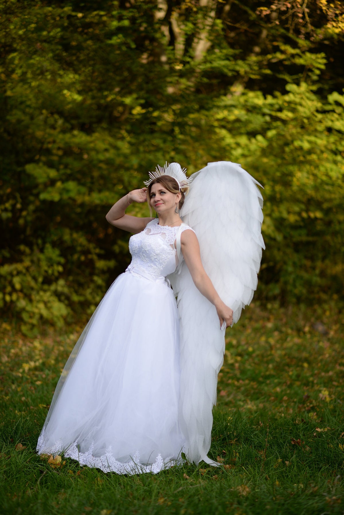 May include: A woman in a white wedding dress with lace detailing and large white angel wings stands in a grassy area. She wears a crown-like headpiece. The background features green foliage.