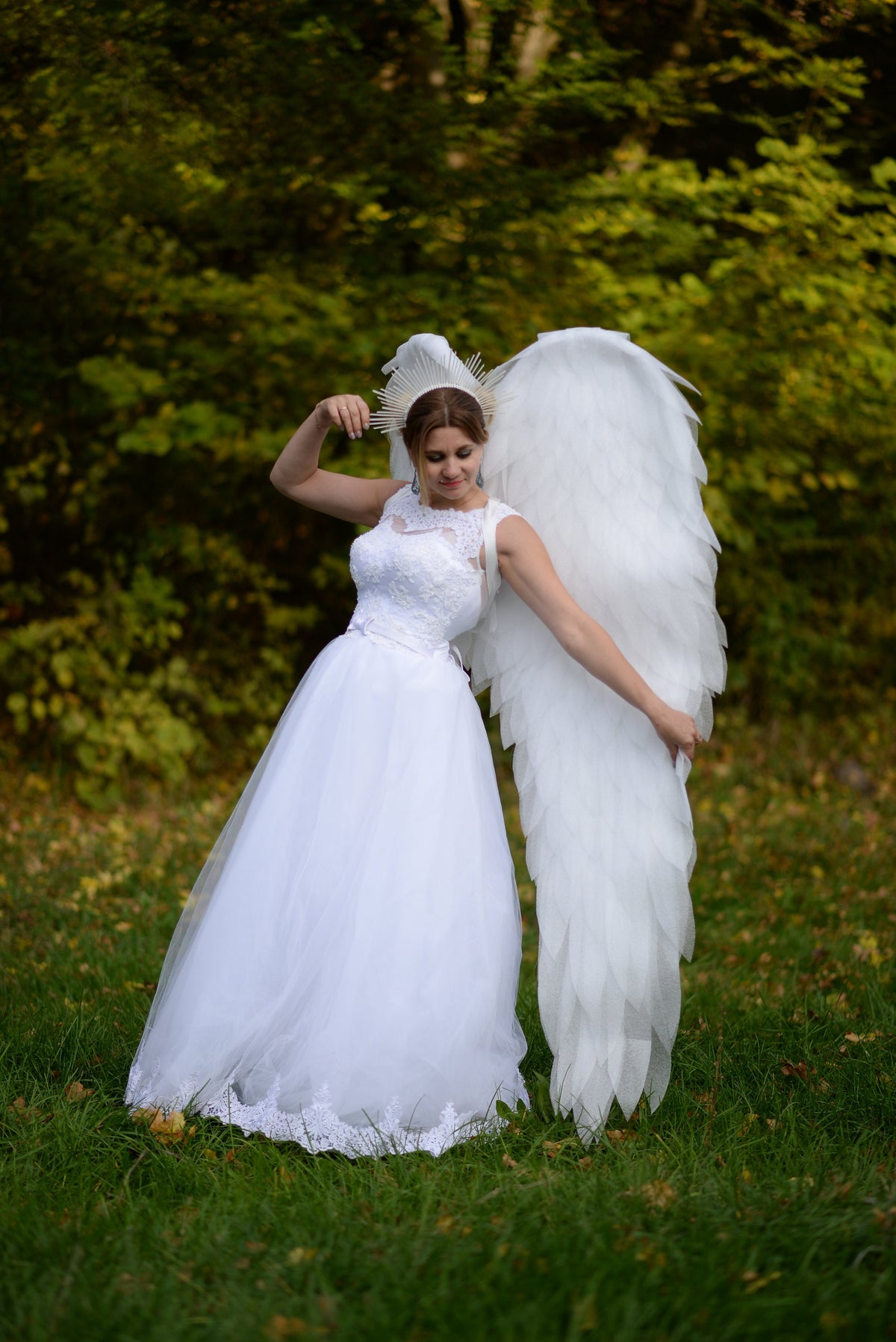 May include: A woman in a white wedding dress with lace details and large white feathered angel wings stands in a grassy area. She wears a halo-like headpiece. The background features green foliage.