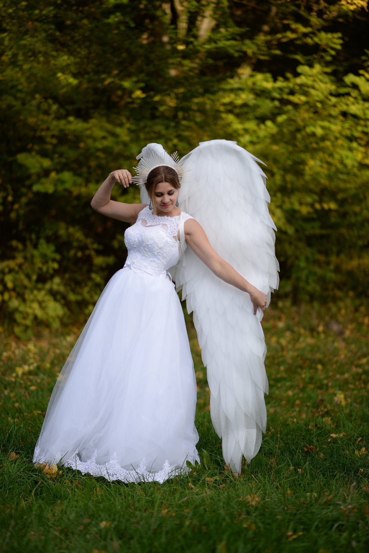 May include: A woman in a white wedding dress with a lace bodice and full skirt, adorned with large white angel wings. She wears a halo-like headpiece. The background is a blurred green forest.