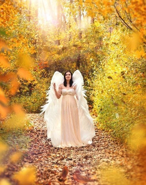 May include: A woman wearing a white dress and angel wings stands in a forest with yellow leaves on the ground.