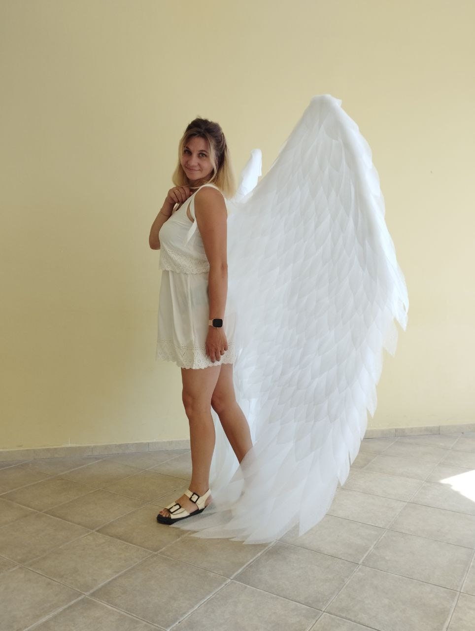 May include: A woman wearing a white dress and white sandals is standing with large white angel wings behind her.