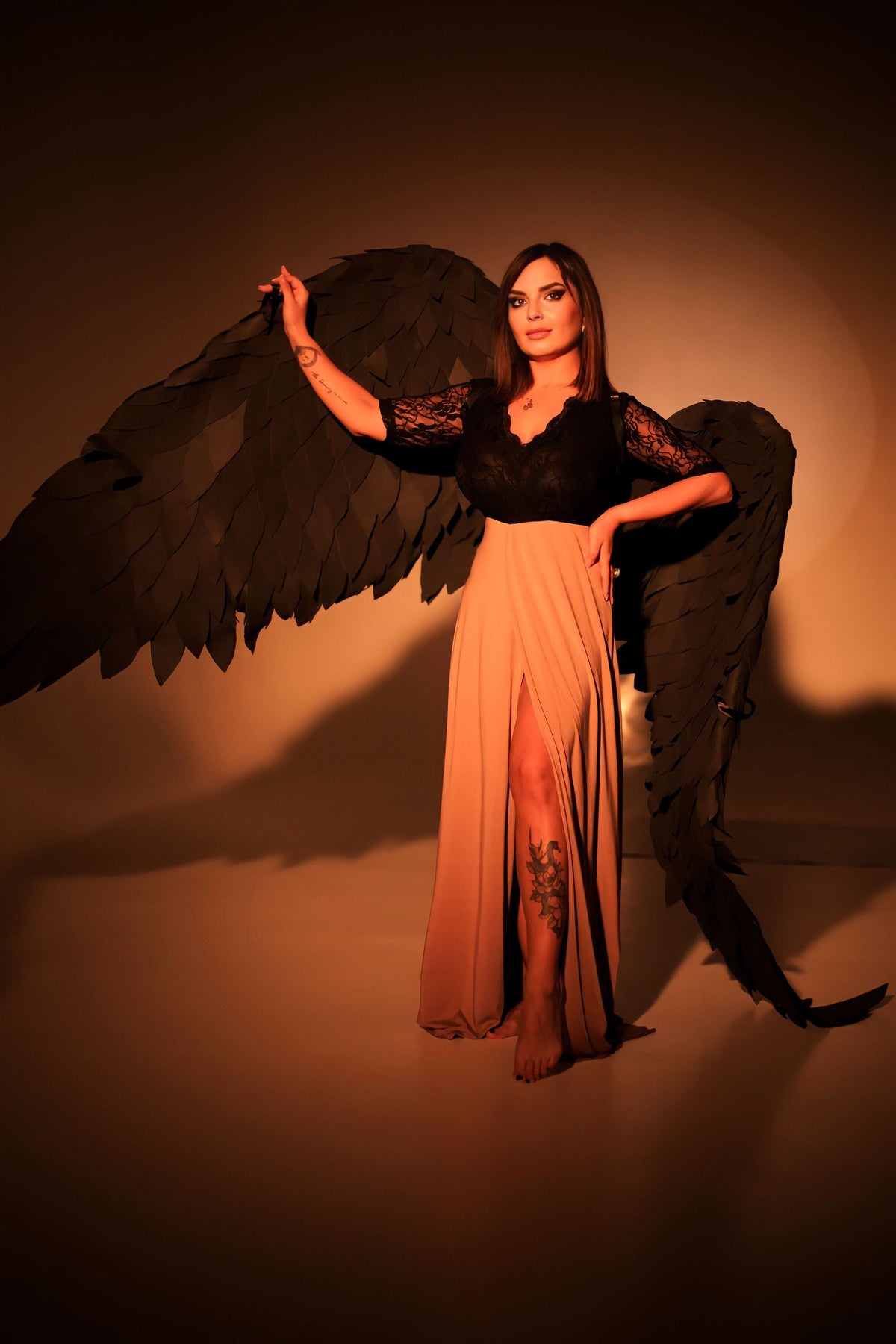 May include: A woman wearing a black lace top and a long, light brown skirt with a slit. She is standing with her arms outstretched, holding large black angel wings.