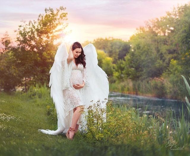 May include: A pregnant woman wearing a white lace dress and large white angel wings stands in a grassy field by a river. The sky behind her is a soft pink and orange.