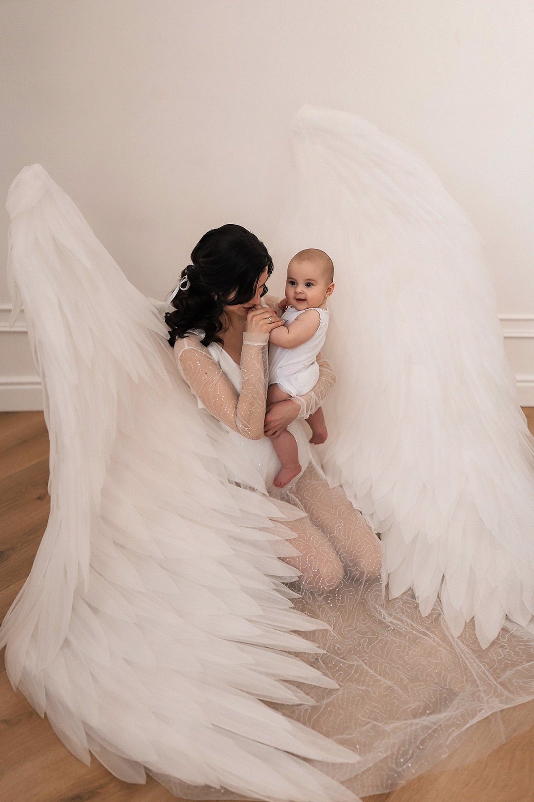May include: A woman and baby are featured in a studio setting. The woman is wearing a white, long-sleeved gown with a feathered skirt and large, white angel wings. The baby is wearing a white onesie. The background is a light color.