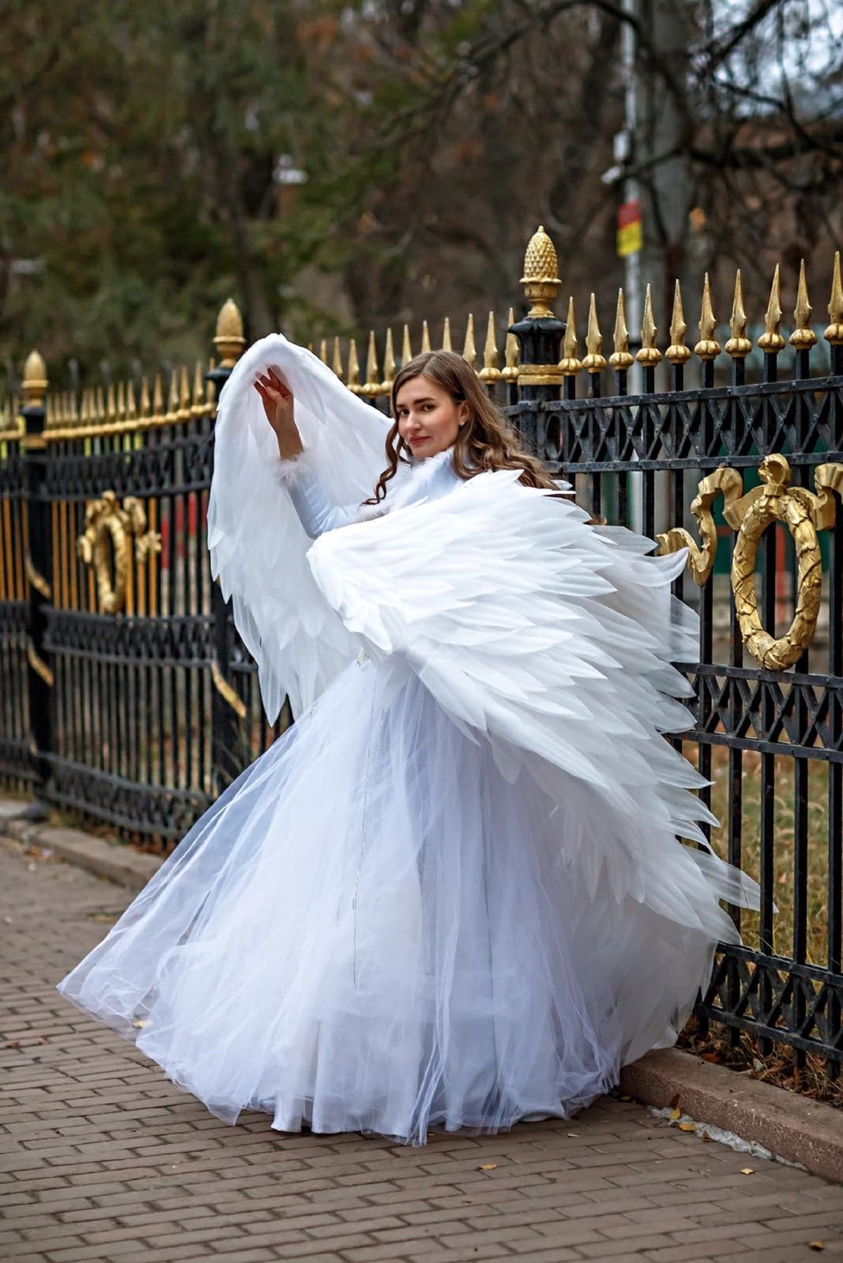 May include: A person wearing a white dress and large white feathered angel wings stands near a black and gold fence. The dress has a long, flowing skirt. The wings are spread wide, creating a dramatic effect.