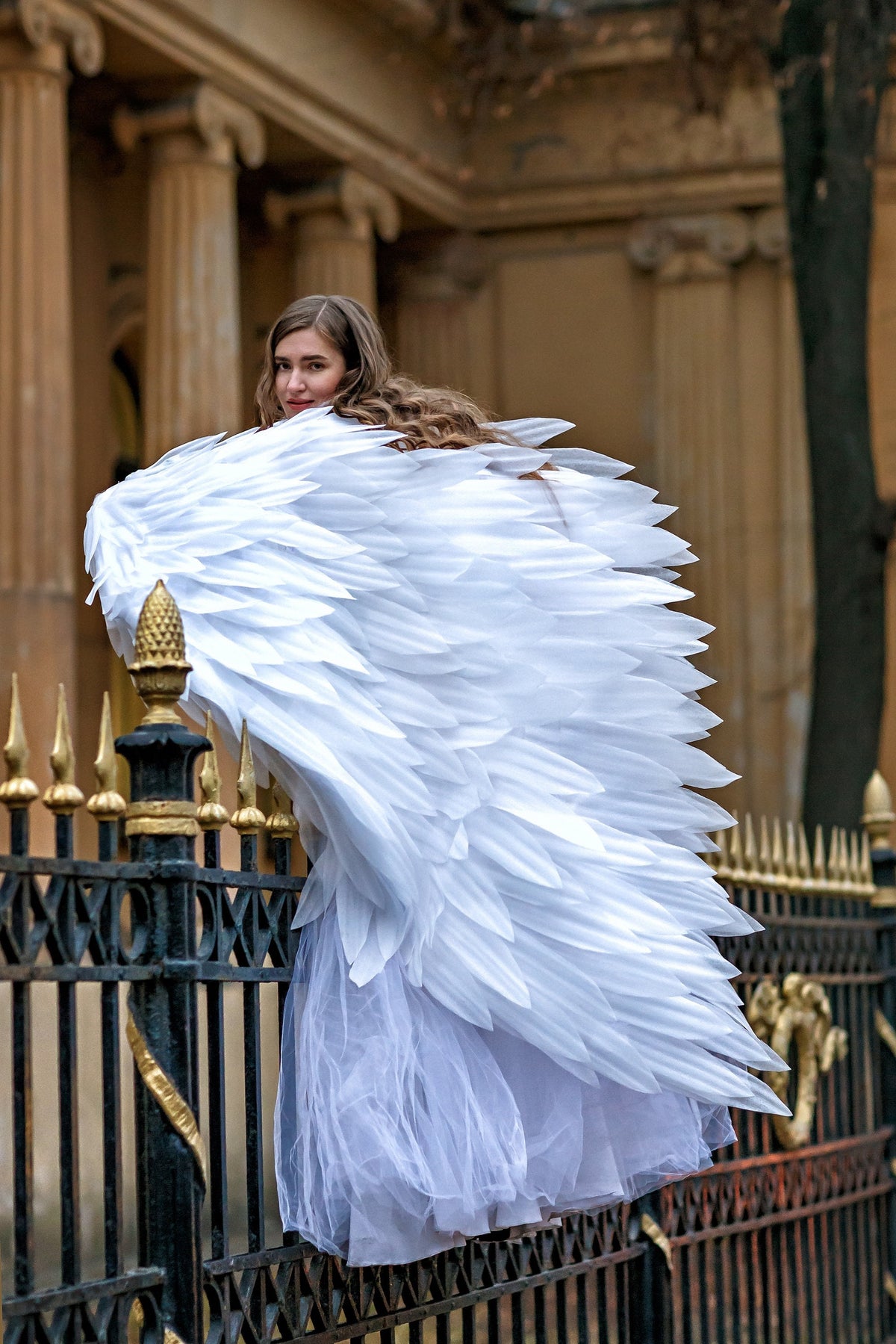 May include: A person wearing large, white feathered angel wings and a white tulle skirt, posing near a black and gold wrought iron fence. The wings are the focal point, with each feather meticulously detailed.