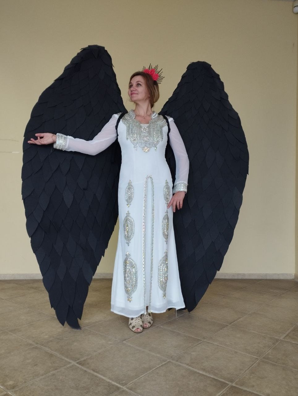 May include: A person wearing a white dress with silver embellishments and a black feather angel wing costume. The wings are large and dramatic, and the person is standing in a room with tiled floors.