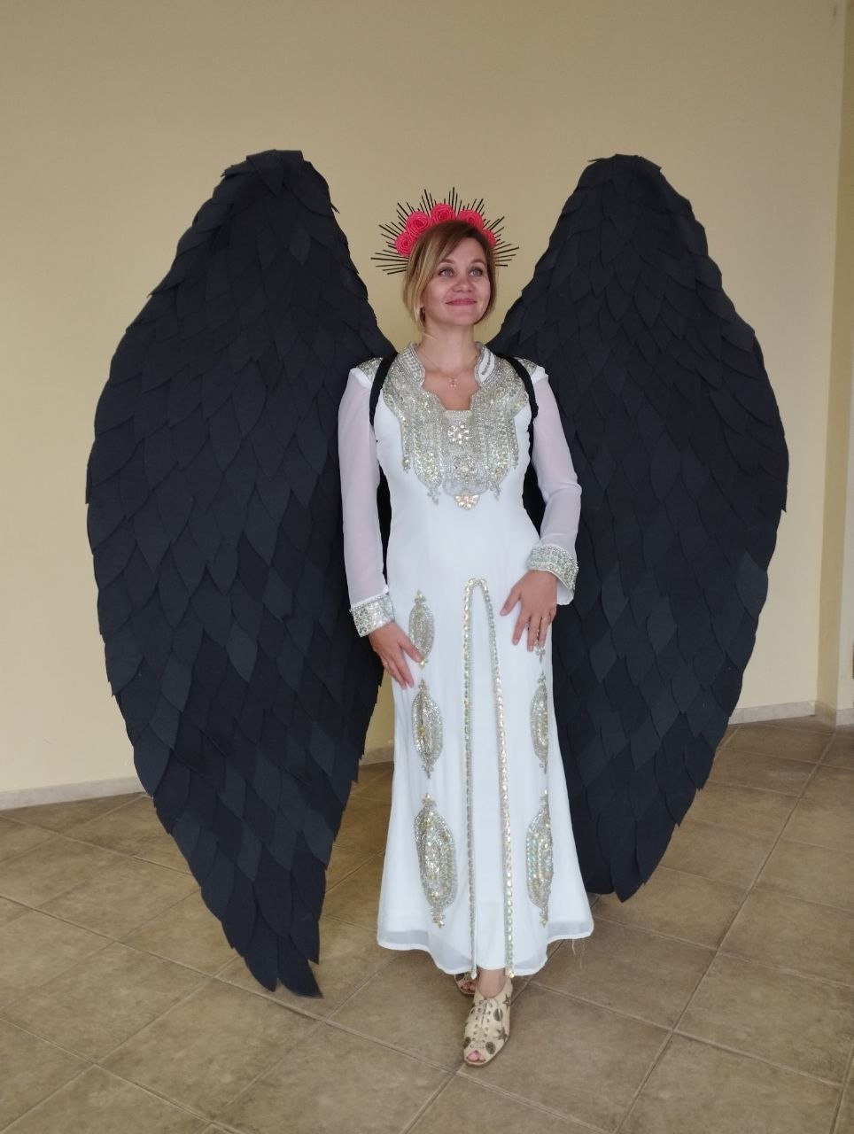 May include: A person wearing a white dress with silver embellishments and a black feather angel wing costume. The person is wearing a flower crown with pink flowers.