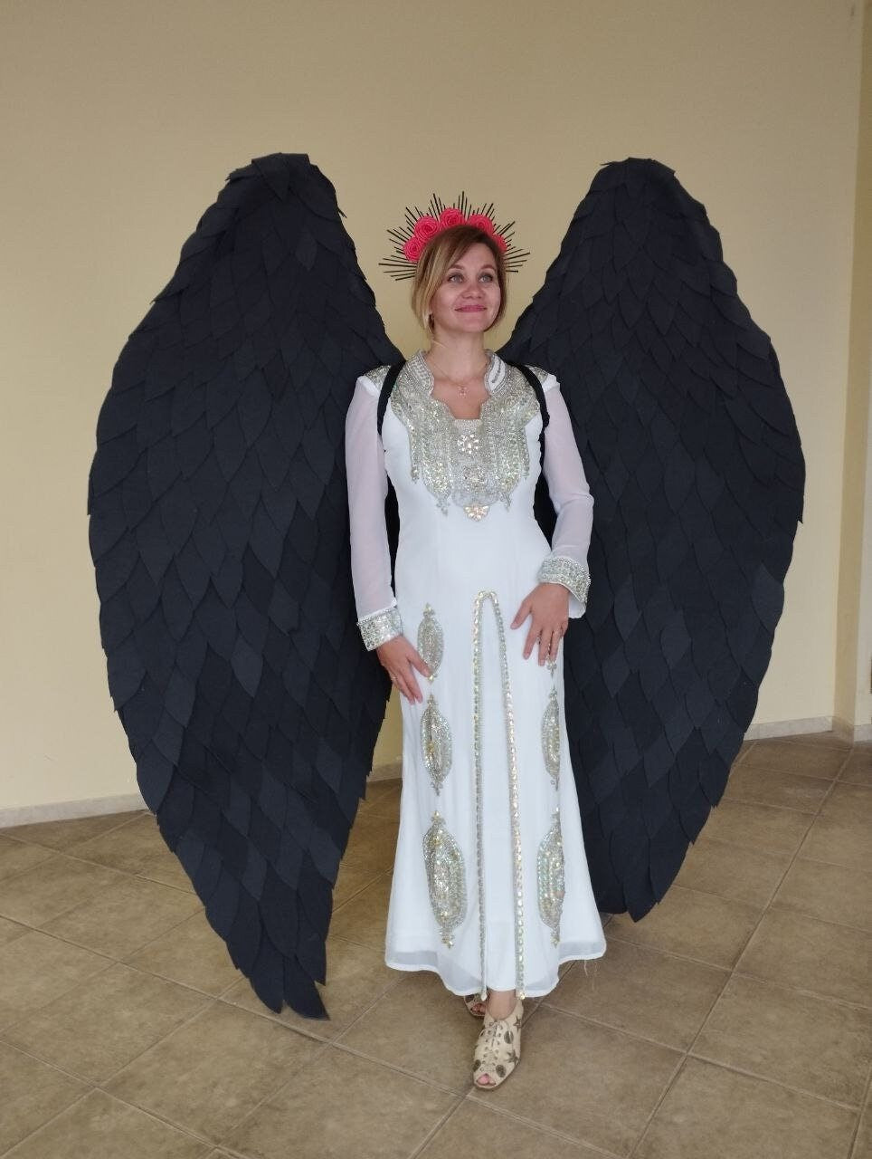 May include: A person wearing a white dress with silver embellishments and a black feather angel wing costume. The person is wearing a flower crown with pink flowers.