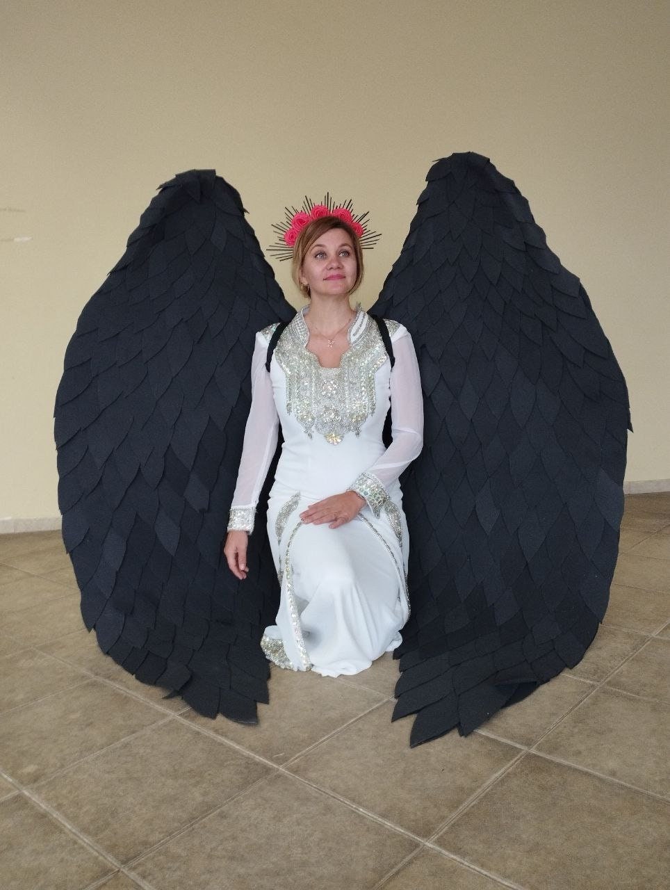 May include: A woman wearing a white dress with a silver design and a flower crown is kneeling with large black wings behind her.