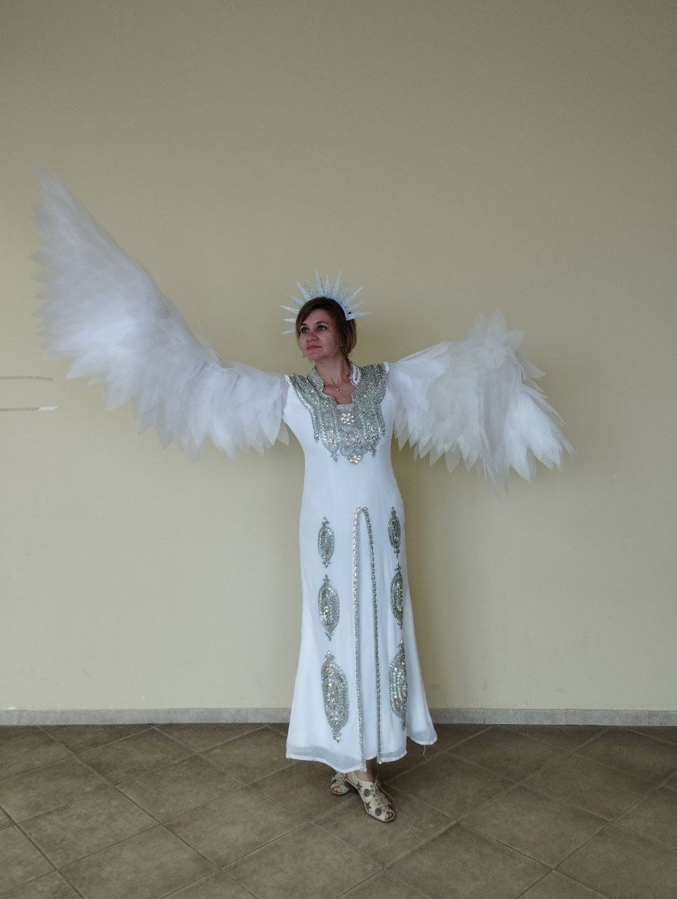May include: A woman wearing a white gown with silver embellishments and a white feathered angel wing costume. She is wearing a silver crown with spikes.