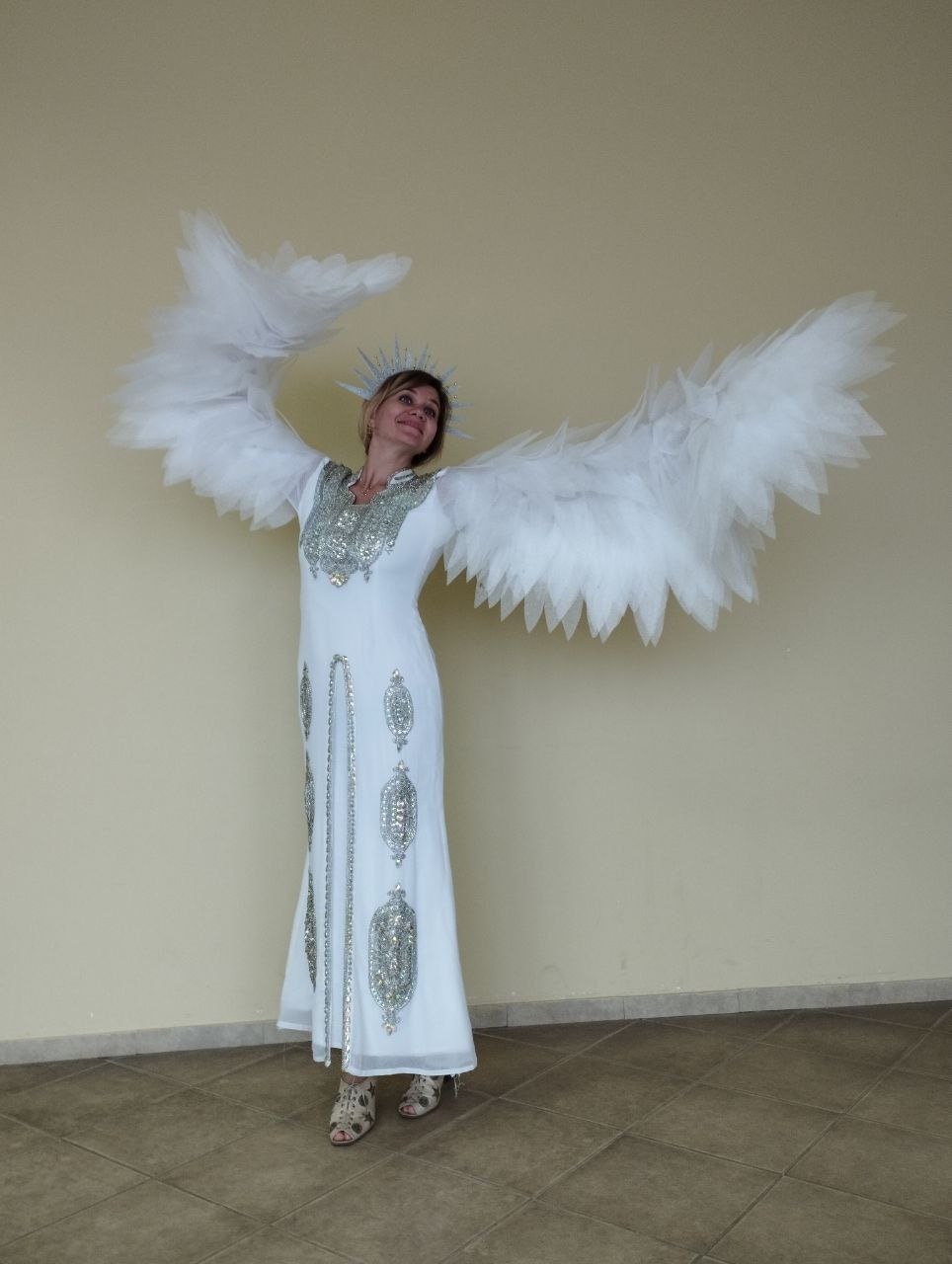 May include: A woman wearing a white dress with silver embellishments and large white feathered wings. She is standing on a tiled floor and looking up.