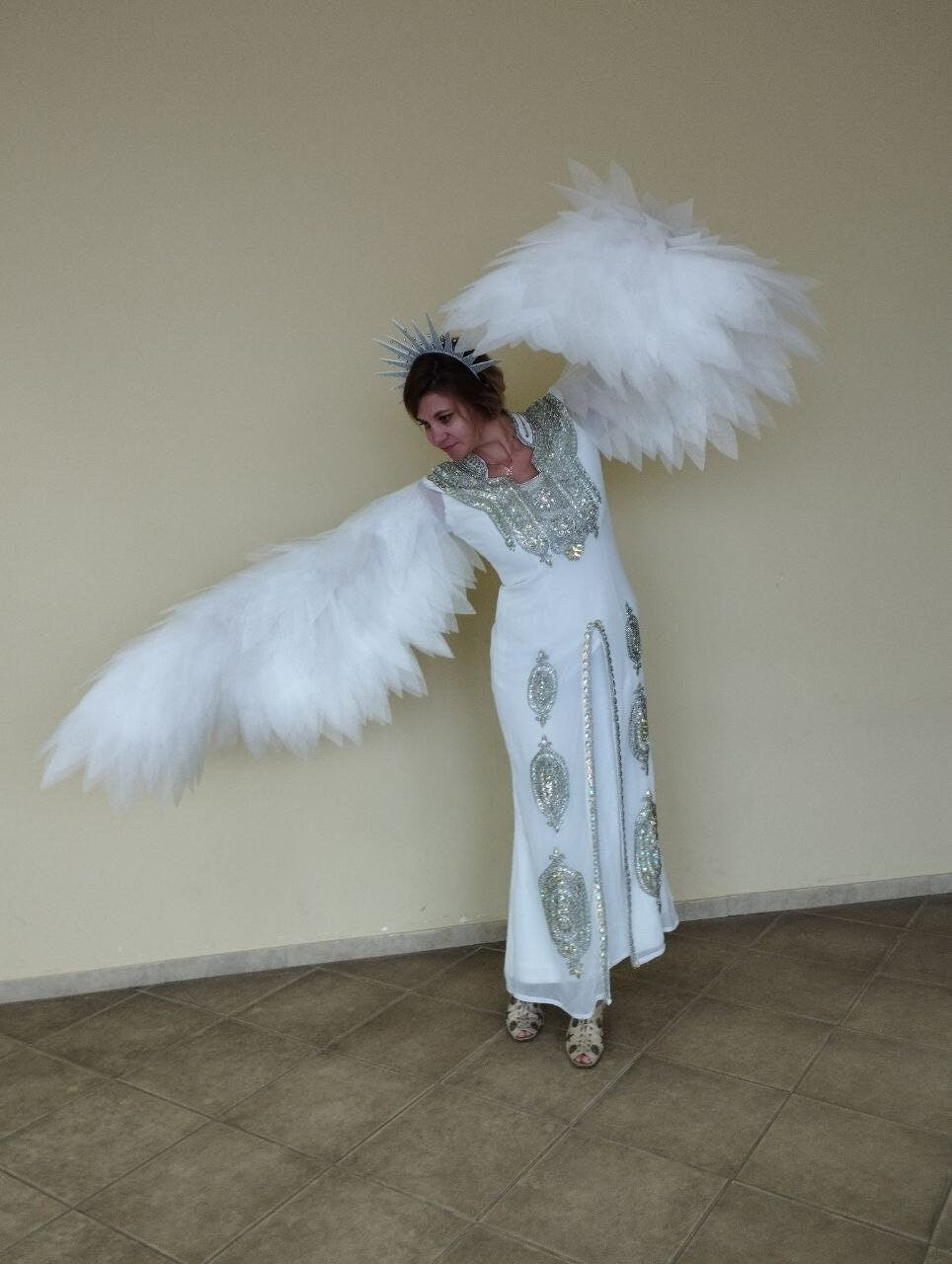 May include: A woman wearing a white dress with silver embellishments and a silver crown. She is holding large white feathered wings out to the sides.