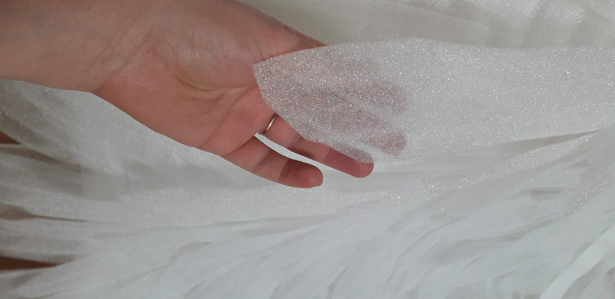 May include: White tulle fabric with a subtle sparkle. The fabric is soft and sheer, and it has a delicate texture.