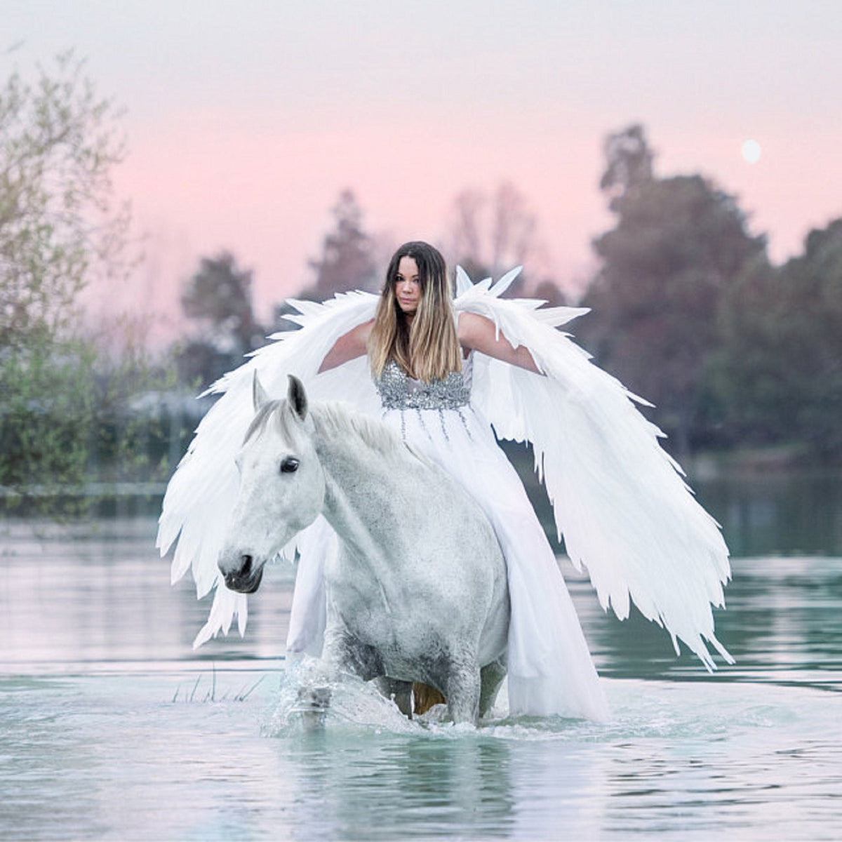 May include: A woman wearing a white dress and large white feathered wings rides a white horse through a lake at sunset.
