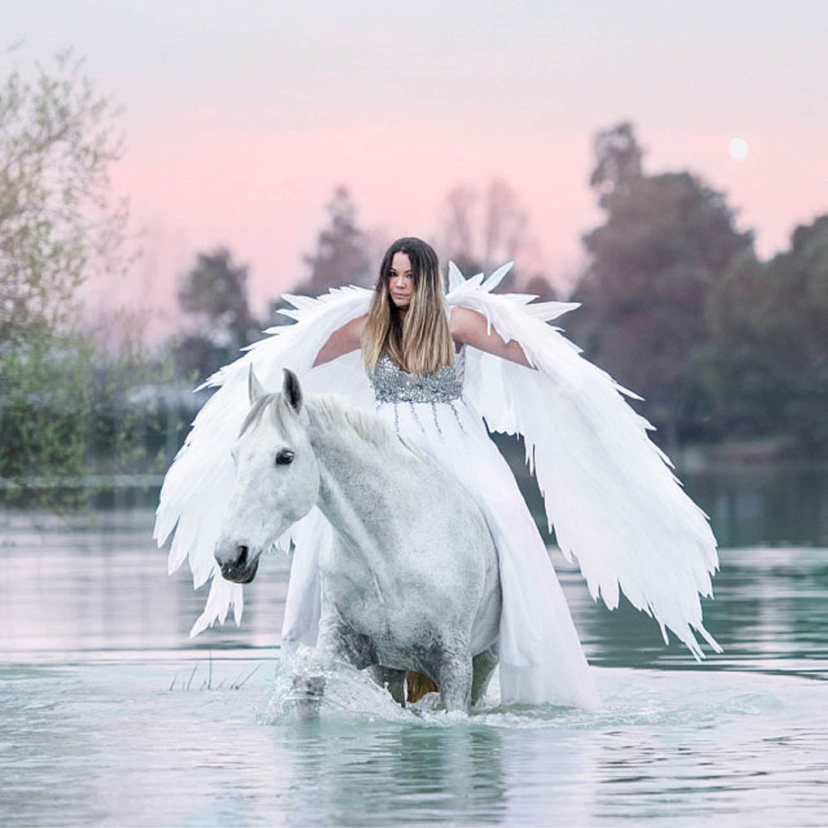 May include: A woman wearing a white dress and large white feathered wings rides a white horse through a lake at sunset.