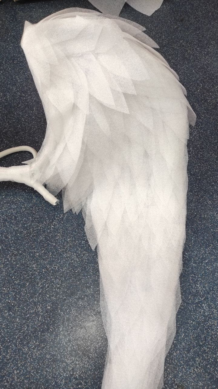 May include: A white, feathery angel wing made of layered fabric. The wing is folded and appears to be part of a costume or prop.