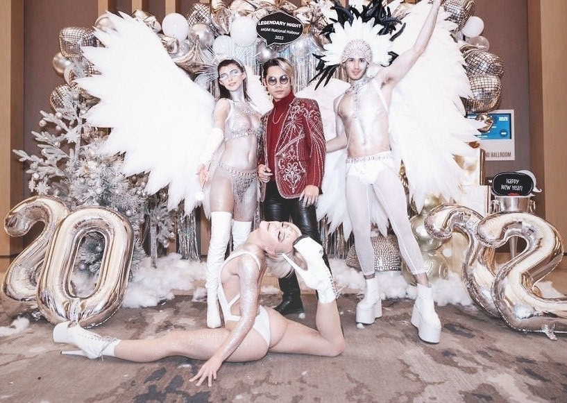 May include: Four people dressed in white and silver costumes pose in front of silver 2022 balloons. The person in the center is wearing a red sequined jacket. The text "LEGENDARY NIGHT" is visible in the background.