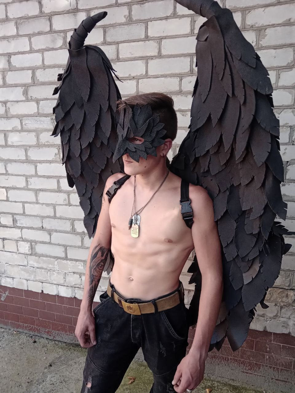 May include: Black costume wings with a matching mask. The wings are large and detailed, with a layered feather design. The mask covers the eyes and is also black. The costume is worn with black pants and a belt.