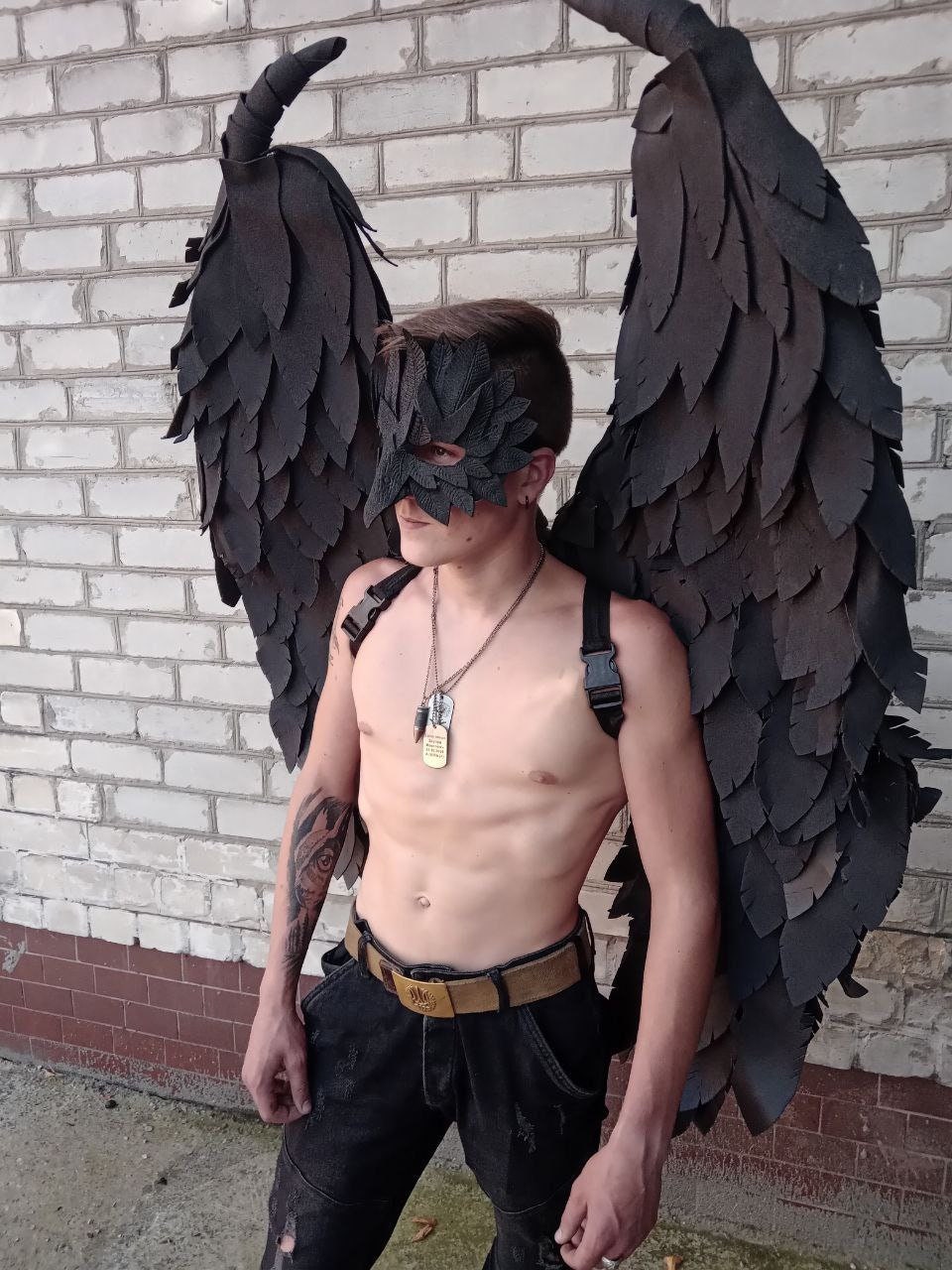 May include: Black costume wings with a matching mask. The wings are large and detailed, with a layered feather design. The mask covers the eyes and is also black. The costume is worn with black pants and a belt.