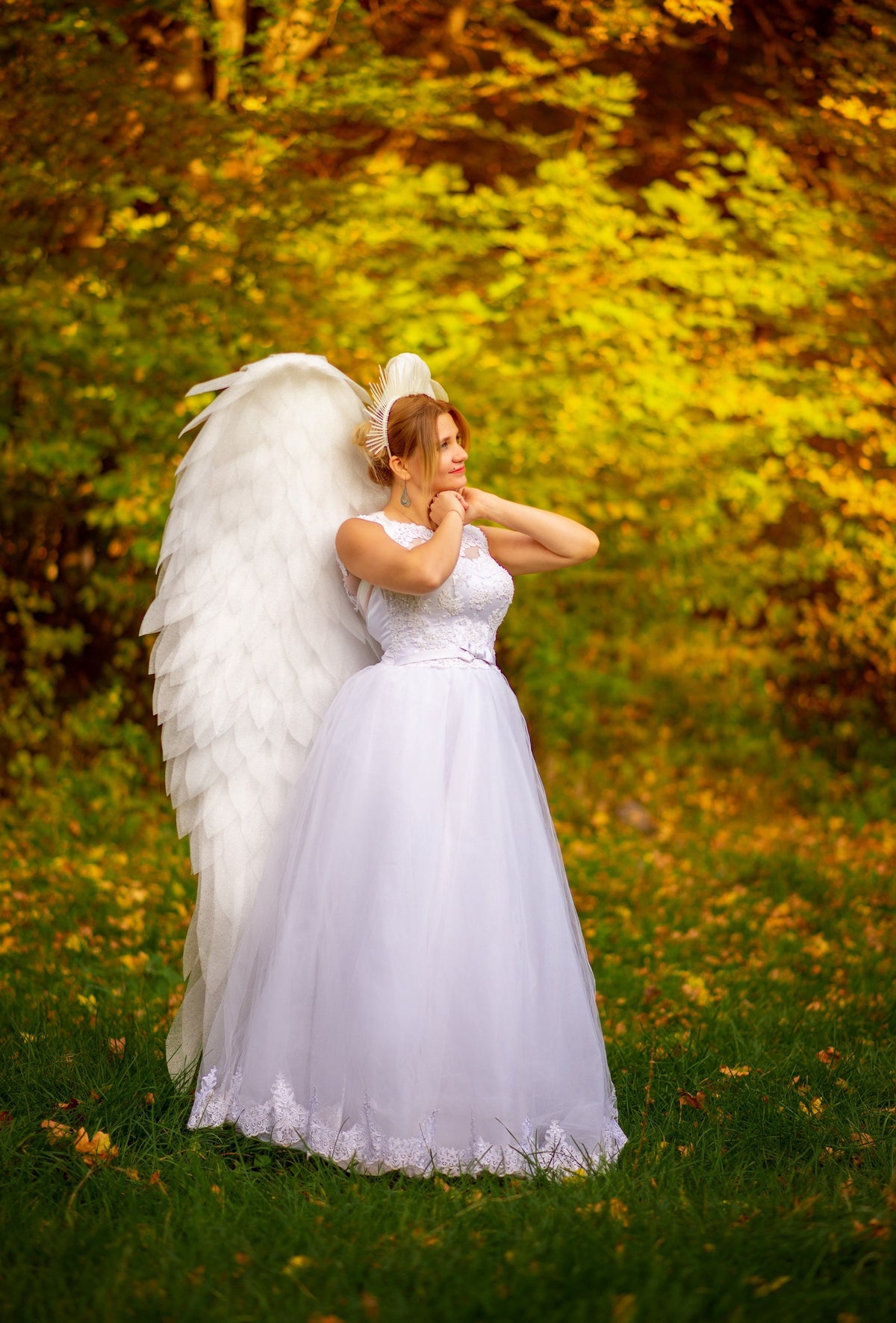 May include: A woman in a white dress with large white angel wings stands in a grassy area with a backdrop of trees. The dress has a fitted bodice and a full, flowing skirt. She wears a headpiece and has her hands clasped near her neck.