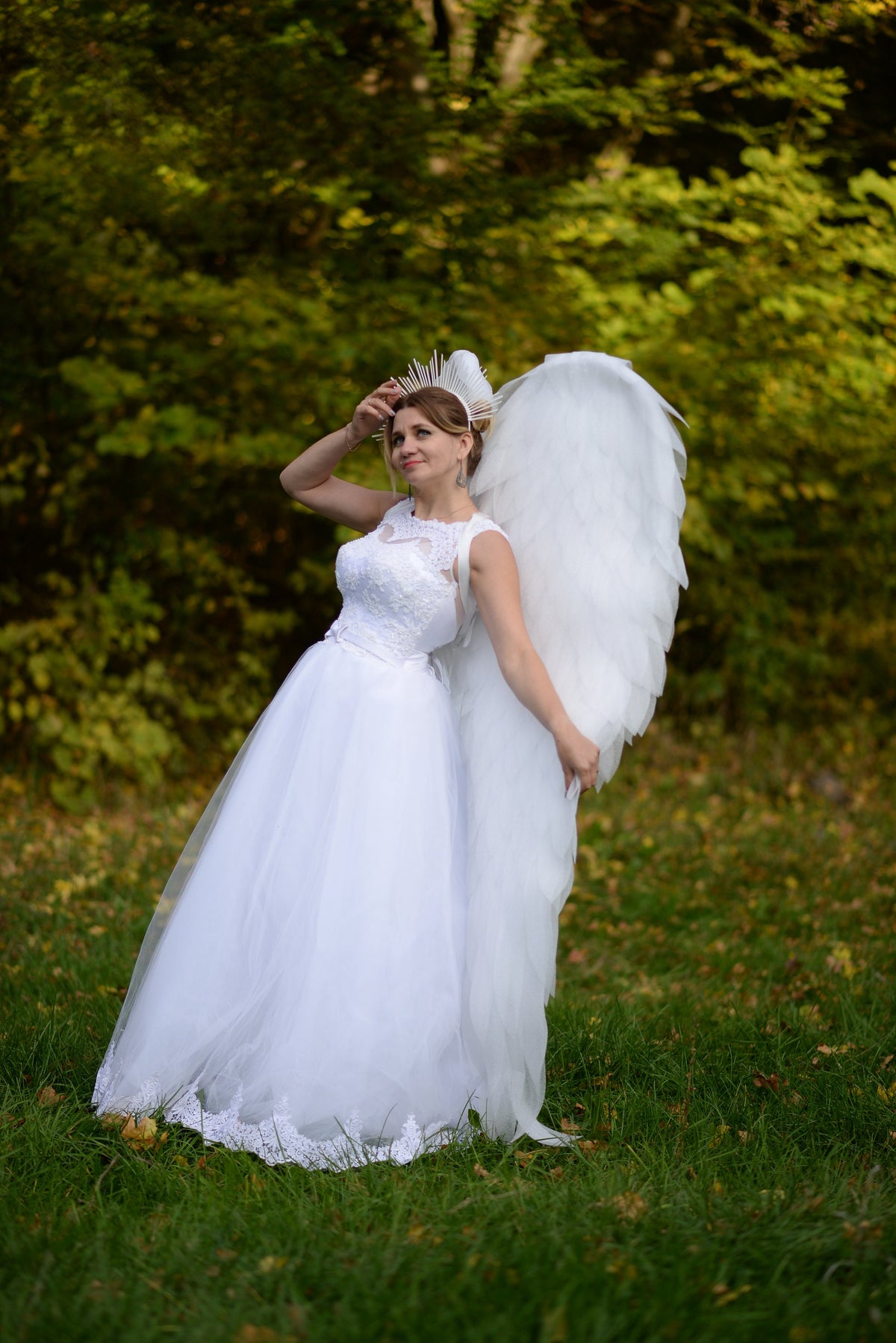 May include: A woman in a white wedding dress with a lace bodice and a full tulle skirt, posing outdoors. She wears a halo and large white feathered wings. The background is a blurred green forest.