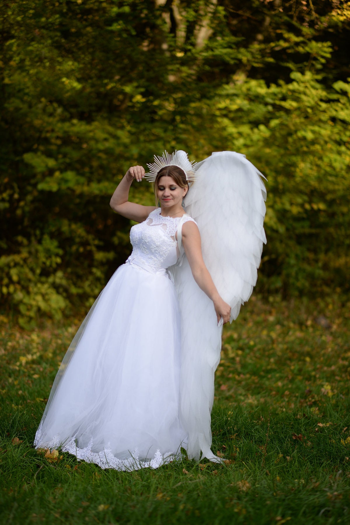 May include: A white wedding dress with a lace bodice and a full tulle skirt. The dress is paired with large white feathered wings and a spiky halo. The person is standing in a grassy area with a blurred green background.