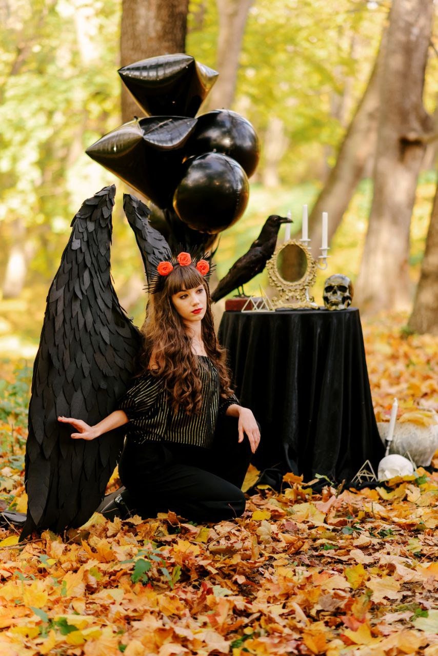 May include: A young woman dressed in black with black feathered wings and a red flower crown sits in a forest setting. She is surrounded by black balloons and a table with a black tablecloth, a raven, candles, and a mirror. The ground is covered in fallen autumn leaves.