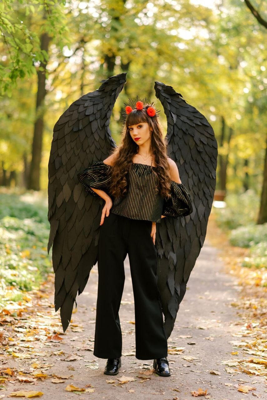 May include: A woman wearing a black and gold striped top and black pants with large black wings. She is wearing a red flower crown.
