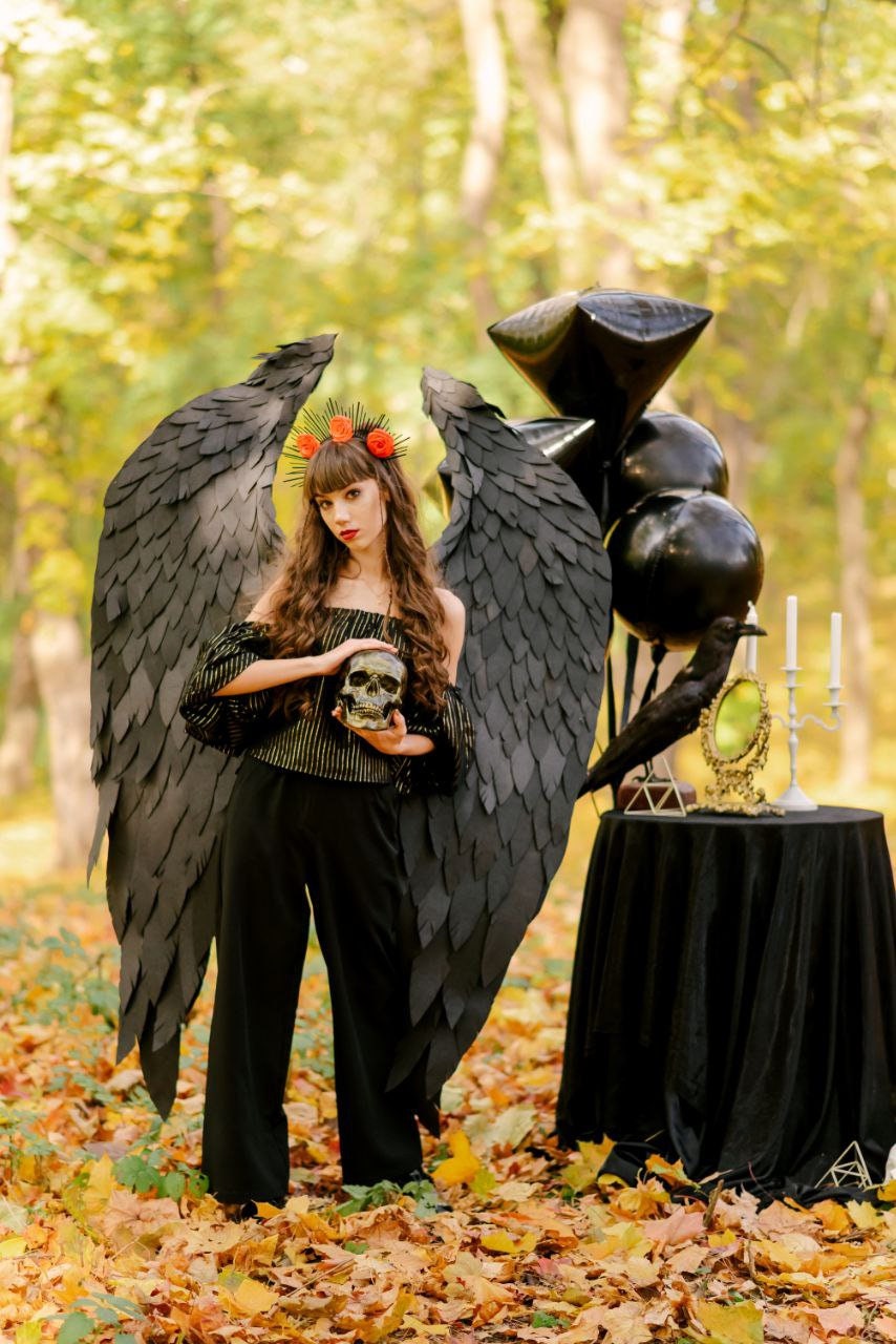 May include: A woman wearing a black costume with large black wings, a skull, and a floral crown stands in a forest setting with autumn leaves on the ground.  Black balloons and a raven are in the background.