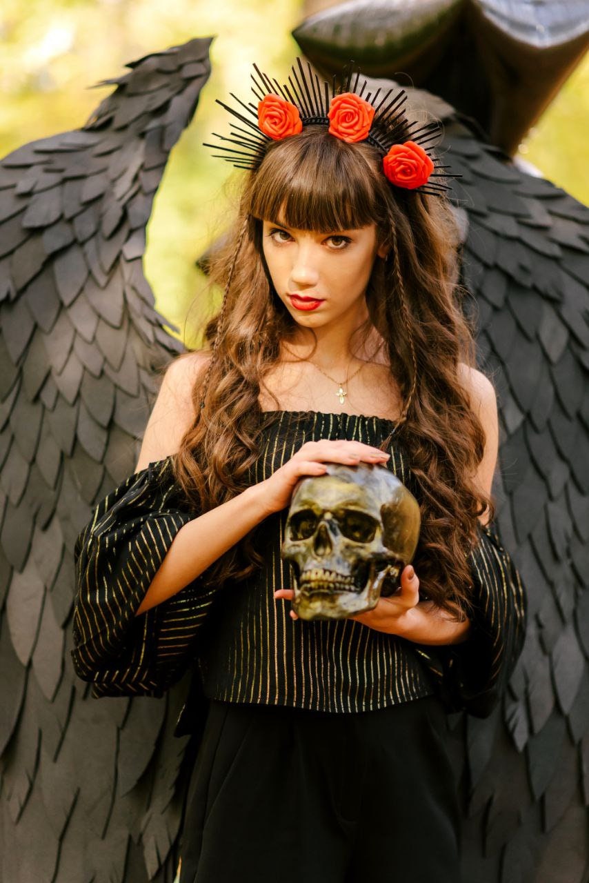 May include: A woman with long brown hair wearing a black and gold striped top and a black skirt. She is holding a skull in her hands. She is wearing a black headband with red flowers. She is standing in front of a black feathered angel wing.