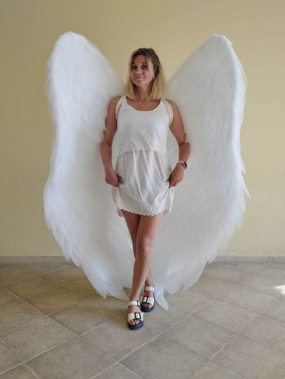 May include: A woman wearing a white dress and white angel wings. The wings are large and feathery, and the woman is standing on a tiled floor.