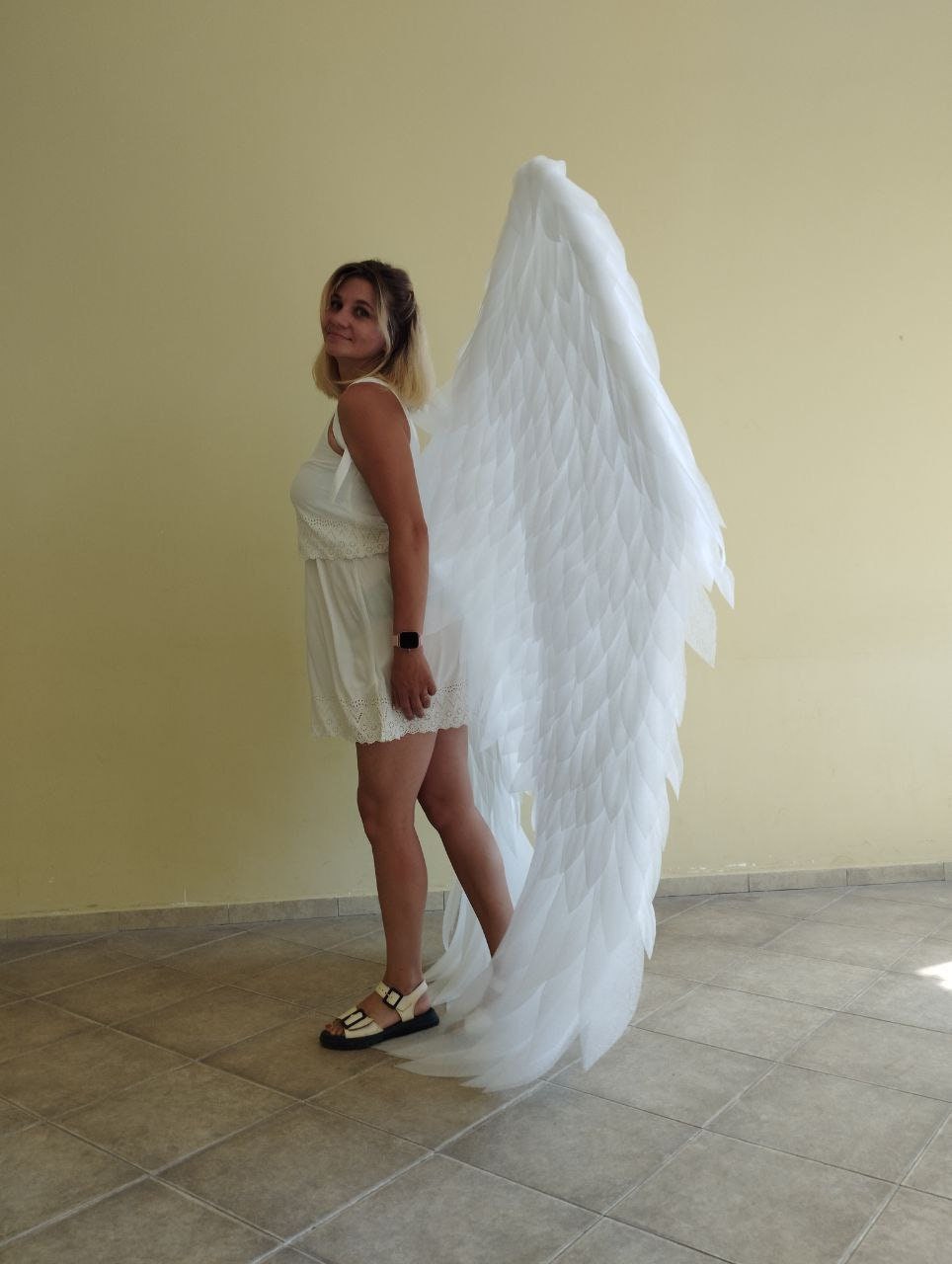 May include: A person wearing a white dress and white angel wings. The wings are large and feathery, and the person is standing on a tiled floor.