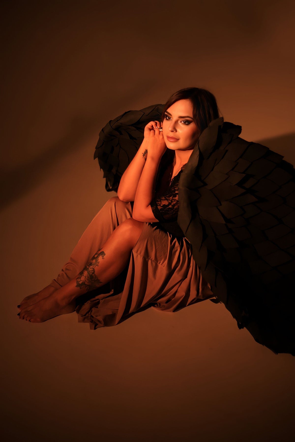 May include: A woman wearing a brown dress and black feathered wings sits with her legs crossed. She has a tattoo on her leg and is looking to the side.
