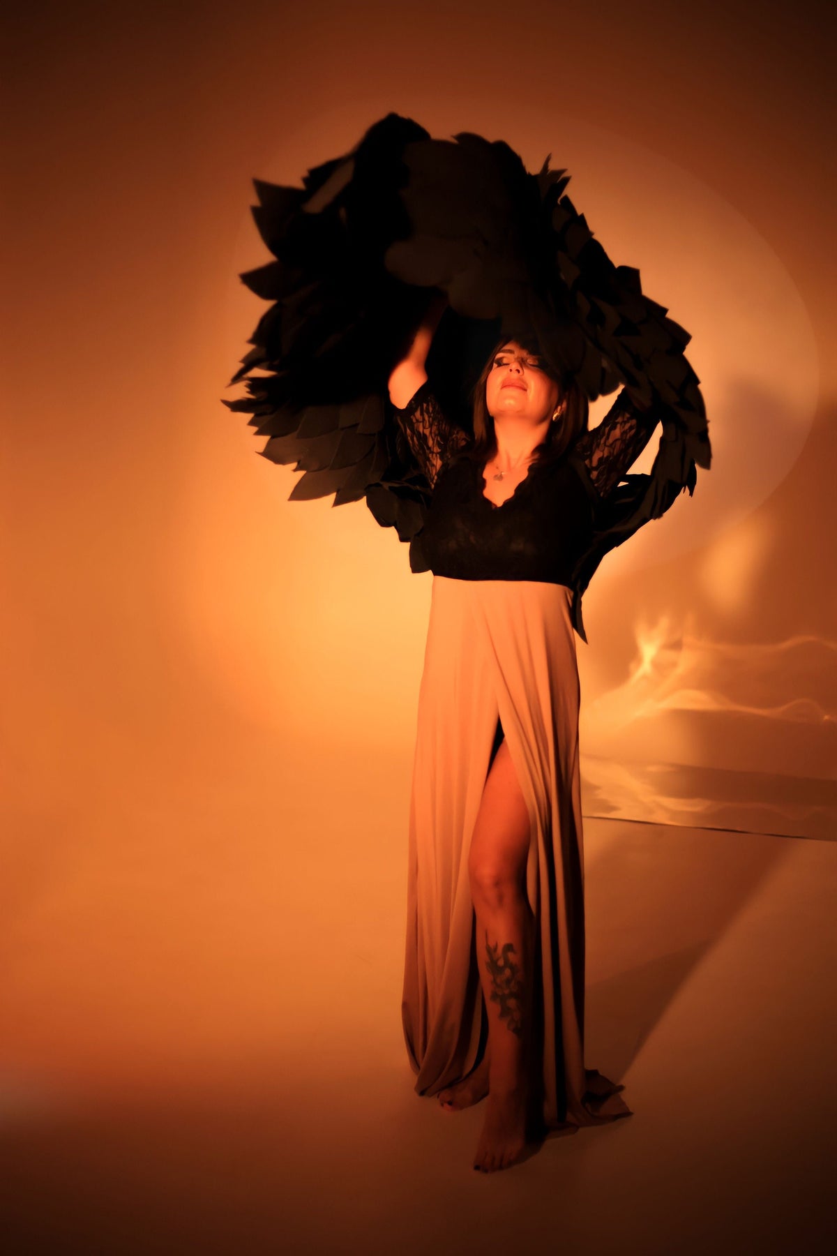 May include: A woman wearing a long, beige dress with a black lace top and a black feathered headdress. The dress has a slit up the side, revealing a tattoo on her leg. The woman is standing in a studio setting with a warm, orange background.