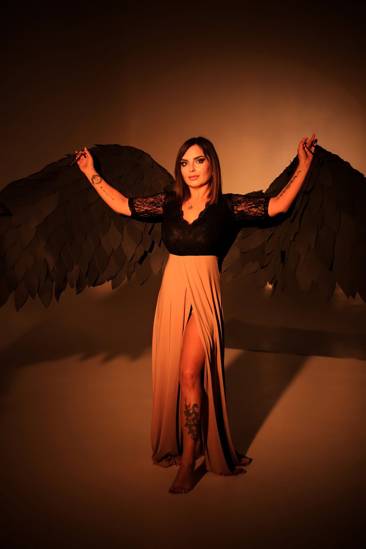 May include: A woman wearing a black lace top and a long beige skirt with a slit. She is holding out large black wings behind her.