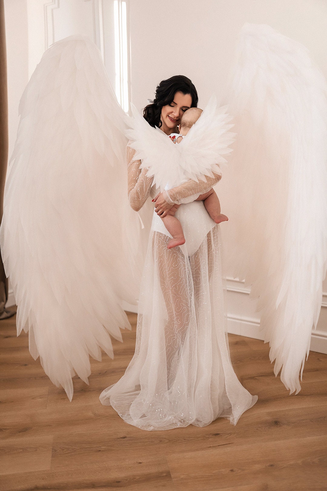 May include: A woman and a baby, both adorned with white angel wings, stand in a well-lit room. The woman wears a sheer, flowing gown, while the baby is held close, also wearing small wings. The scene evokes a sense of purity and innocence.