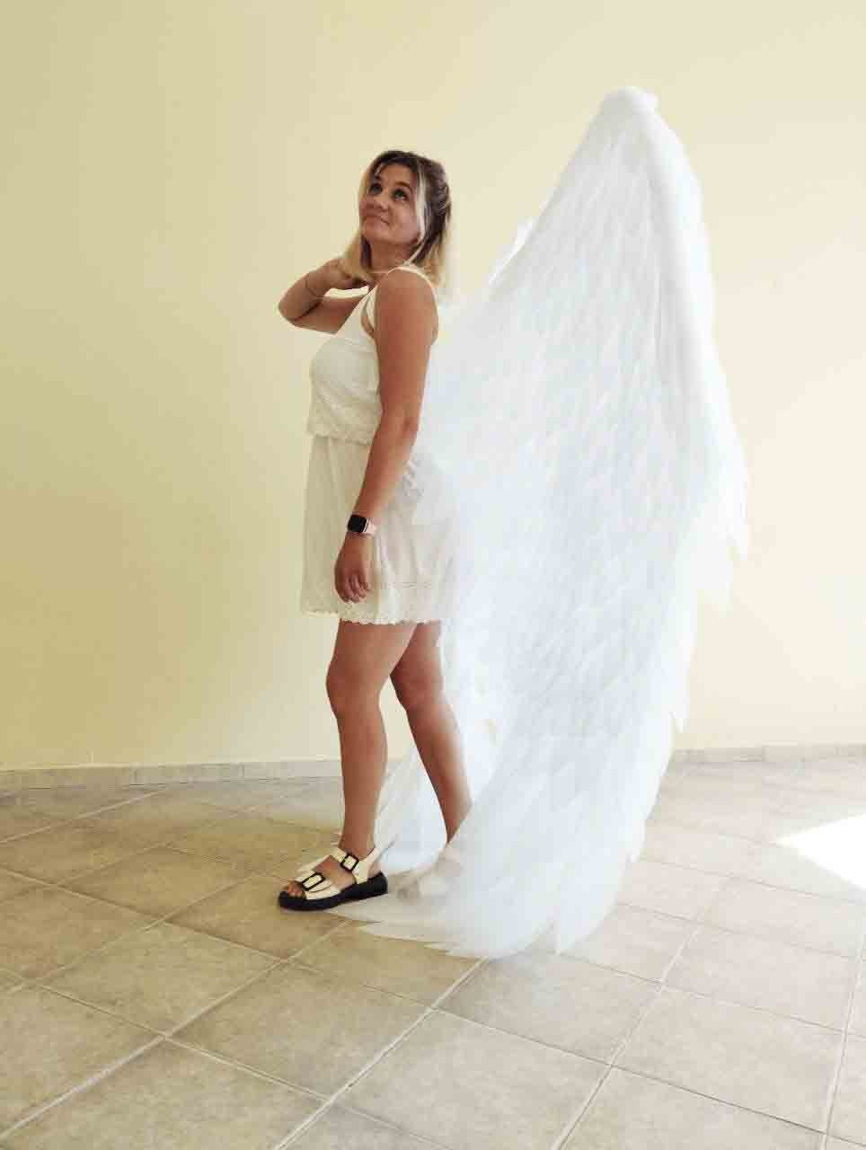 May include: A woman wearing a white dress and sandals stands with large, white feathered angel wings. The wings extend from the shoulders and trail on the ground. The woman is looking up, with her hand on her neck.