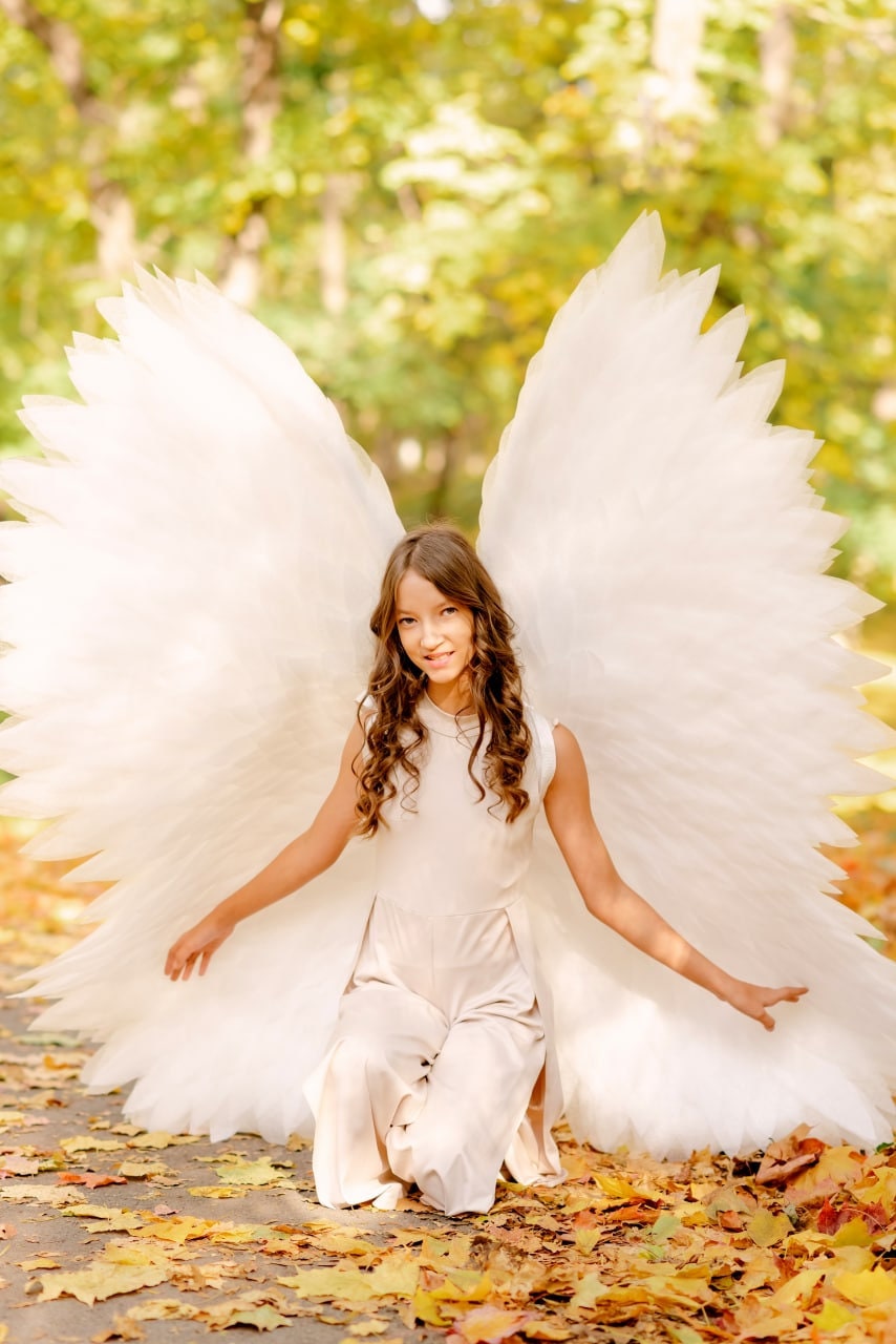 May include: A young person wearing a white dress and large white angel wings, kneeling in a bed of fallen autumn leaves.
